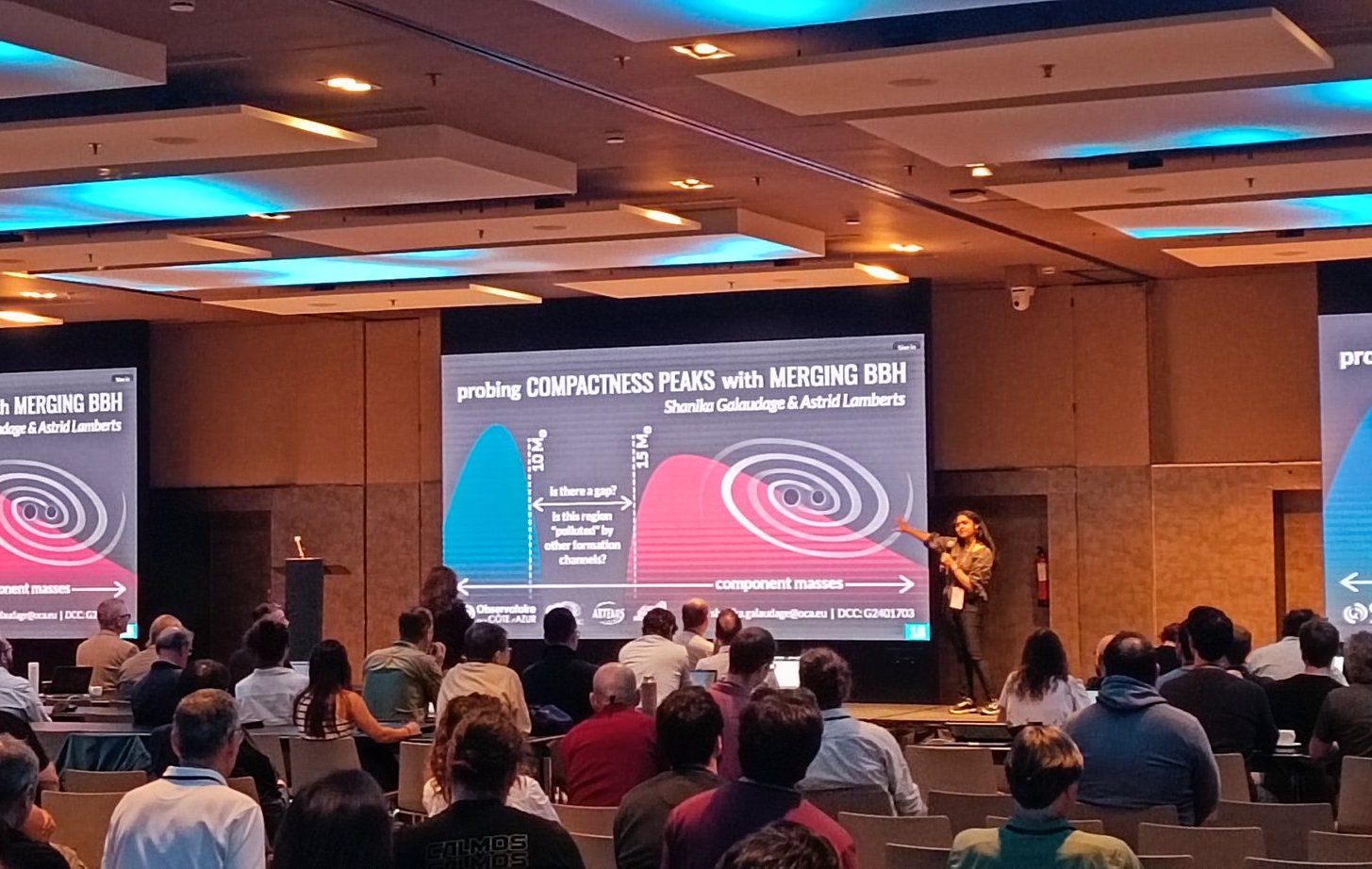 Image of a woman presenting in front of a large projected slide to an audience filling a large hotel conference room.